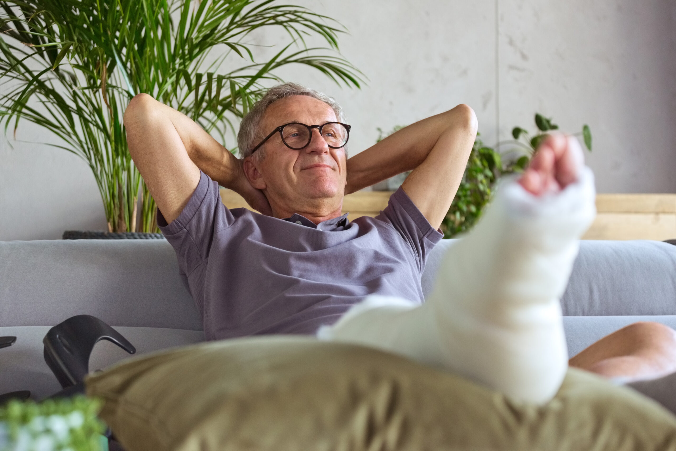 Man ligt op de bank met zijn onderbeen in het gips, ondersteund door een kussen.